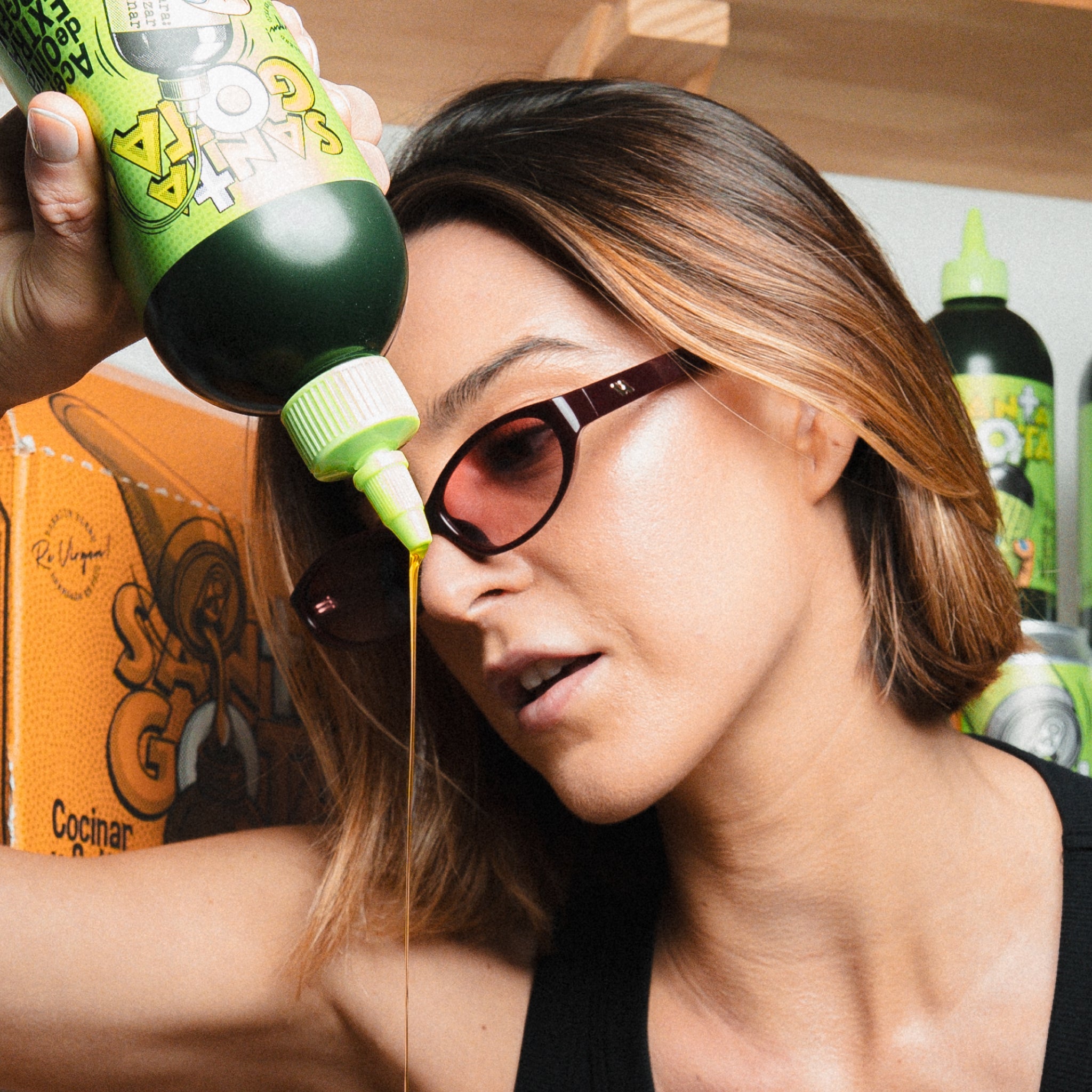 Mujer con gafas de sol vertiendo aceite de oliva desde un squeeze verde Santa Gota, con el packaging al fondo